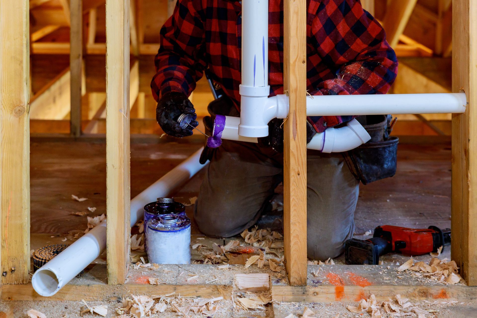 A person in a red plaid shirt kneels between wooden wall studs, installing white PVC pipes with purple primer and tools.