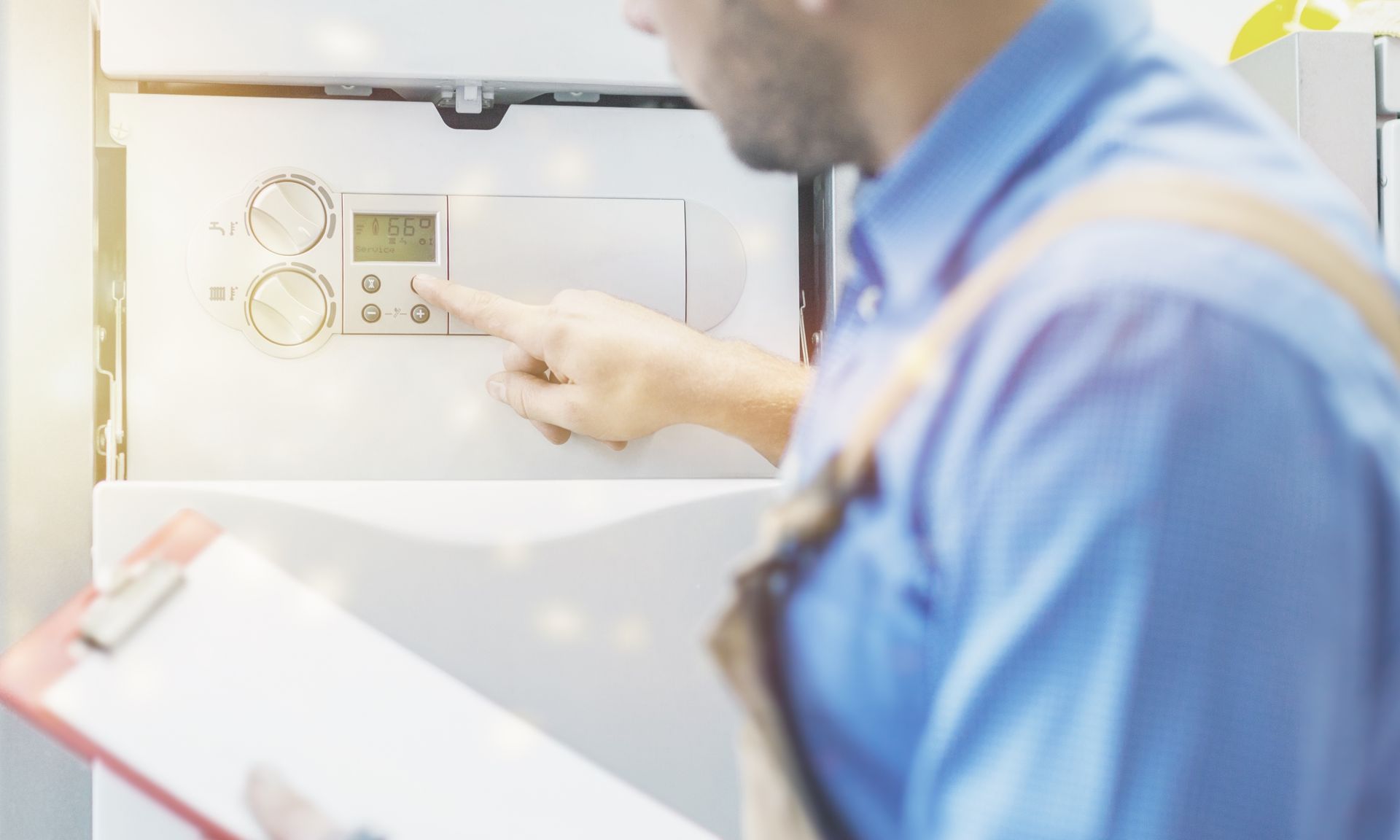 A technician in a blue shirt holds a clipboard while adjusting the settings on a wall-mounted boiler.