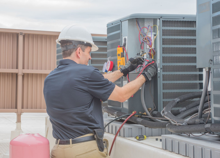 A technician wearing a hard hat and gloves repairs an outdoor HVAC unit on a rooftop.