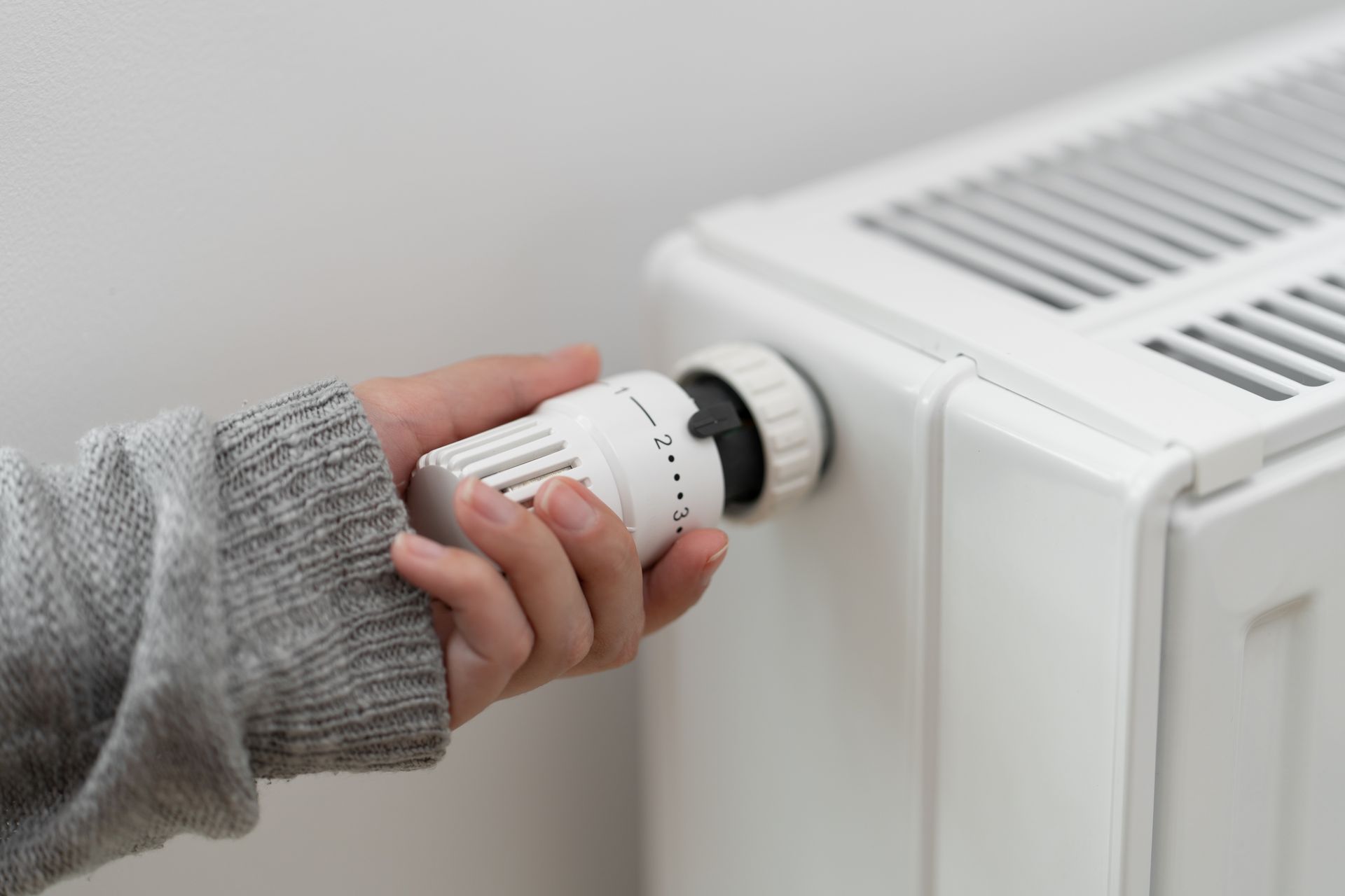 A hand in a grey sweater adjusts the white thermostatic radiator valve on a white radiator.