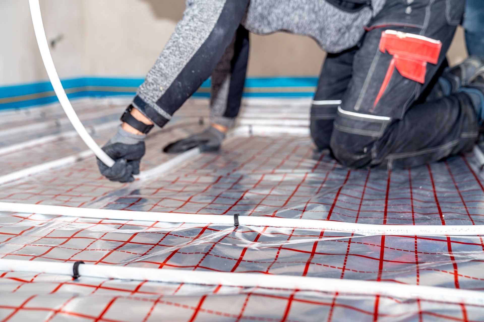 A worker kneels on a floor, installing white tubing over a grid-patterned reflective insulation sheet.