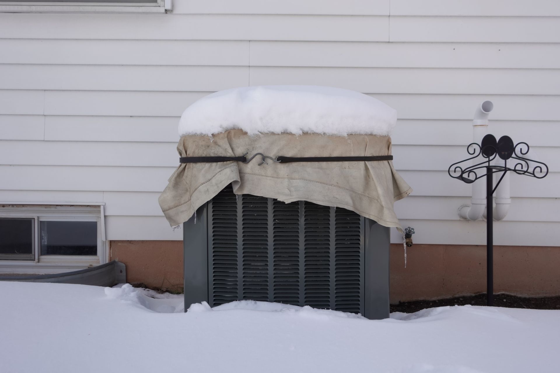 An outdoor air conditioning unit covered with a tan cloth and topped with a layer of snow next to a house.