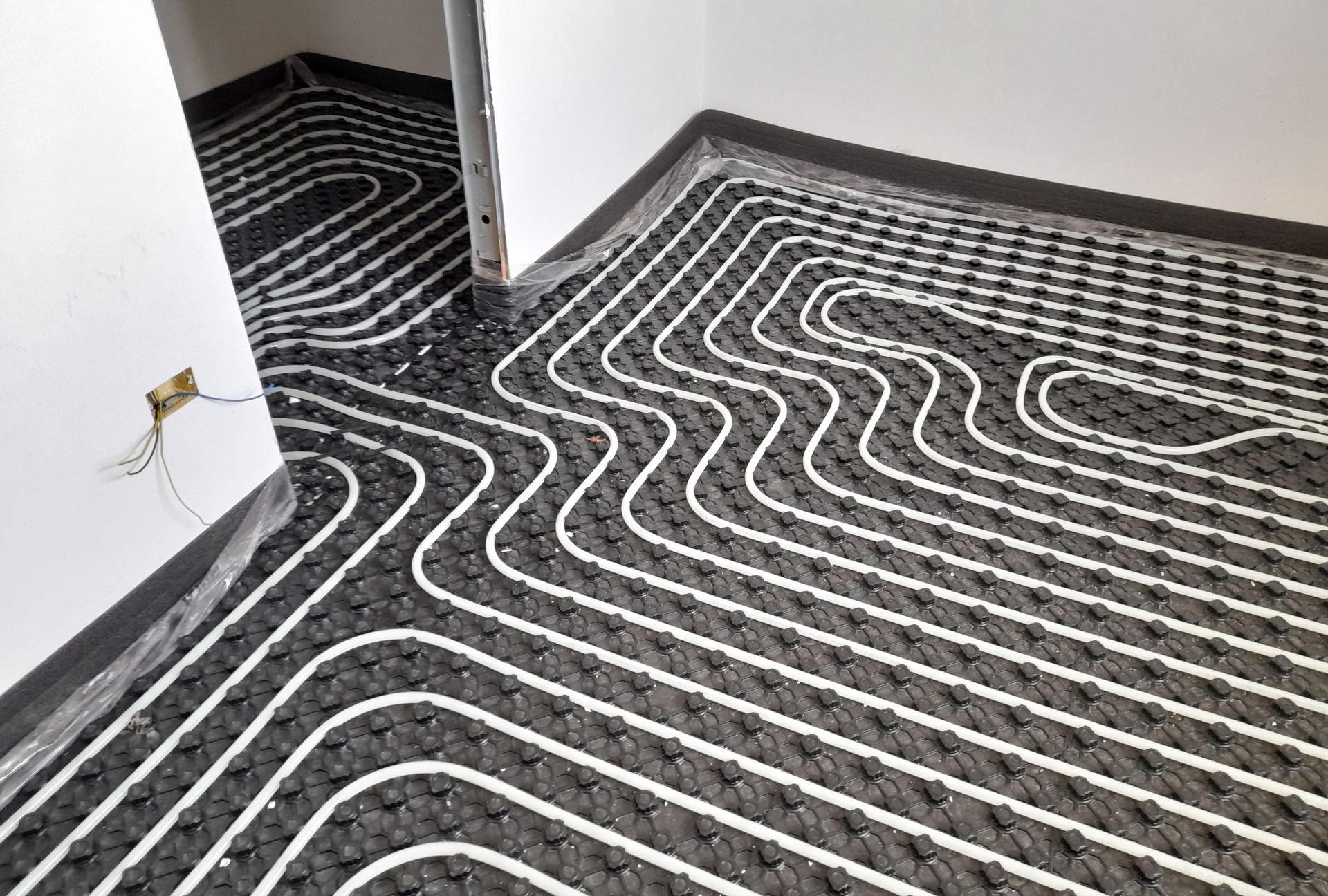 White heating pipes are installed in a serpentine pattern across a grey, dimpled underlayment floor in a room under renovation.
