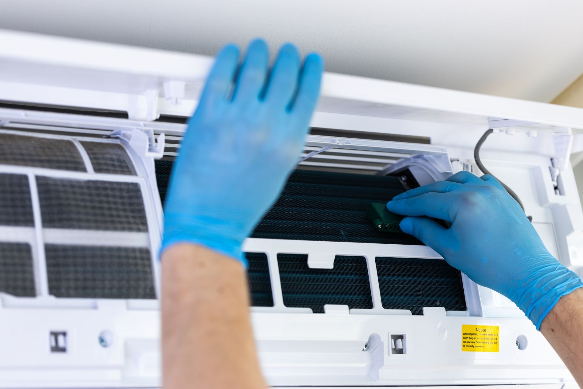 Hands in blue gloves cleaning or inspecting the interior components of a white wall-mounted air conditioner.