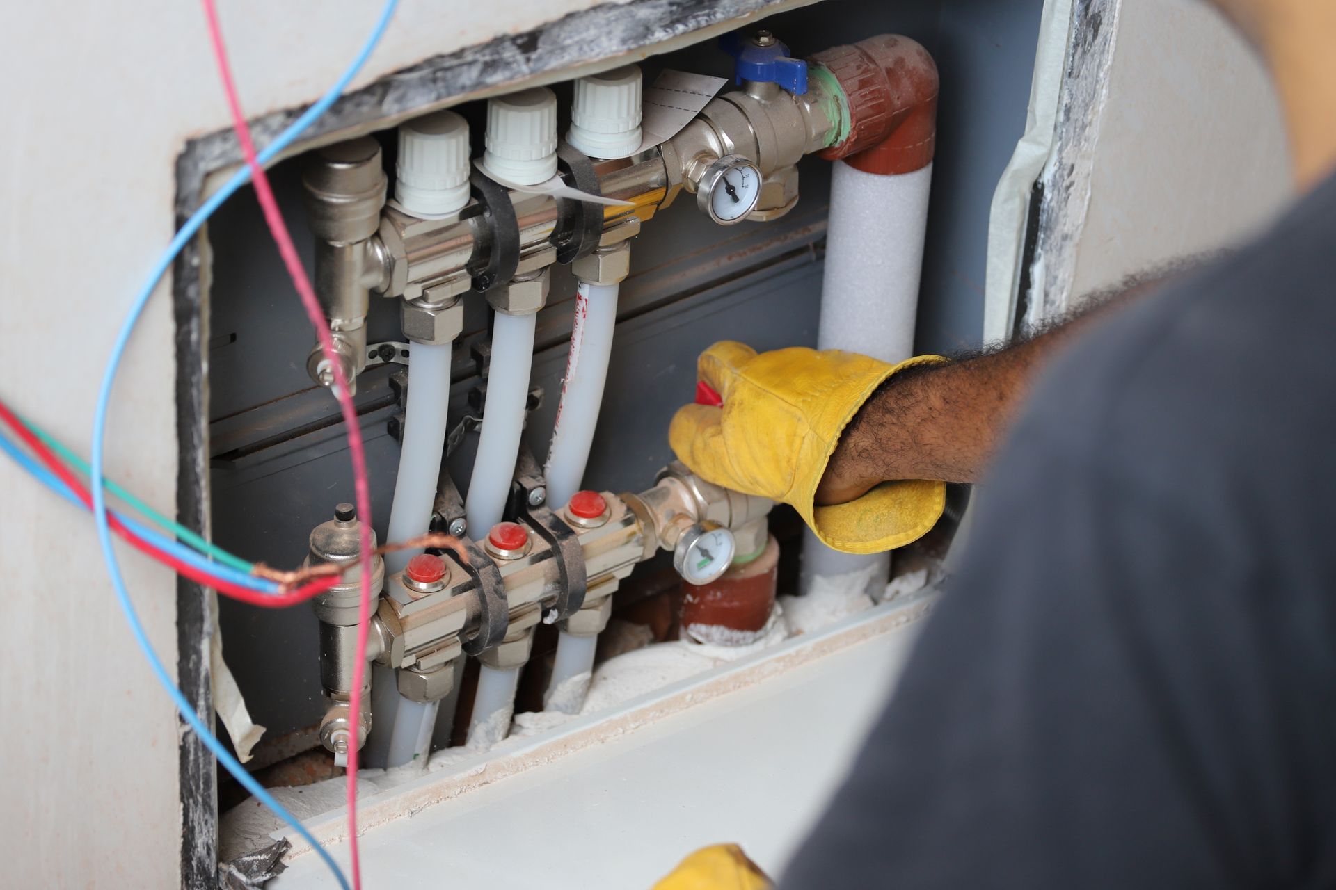 A person wearing work gloves adjusts a plumbing manifold with white plastic pipes inside a wall cavity.