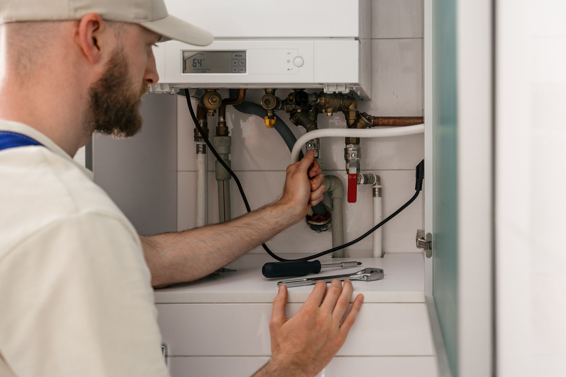 A professional in a white cap and uniform repairs pipes on a wall-mounted gas boiler with tools on the shelf below.
