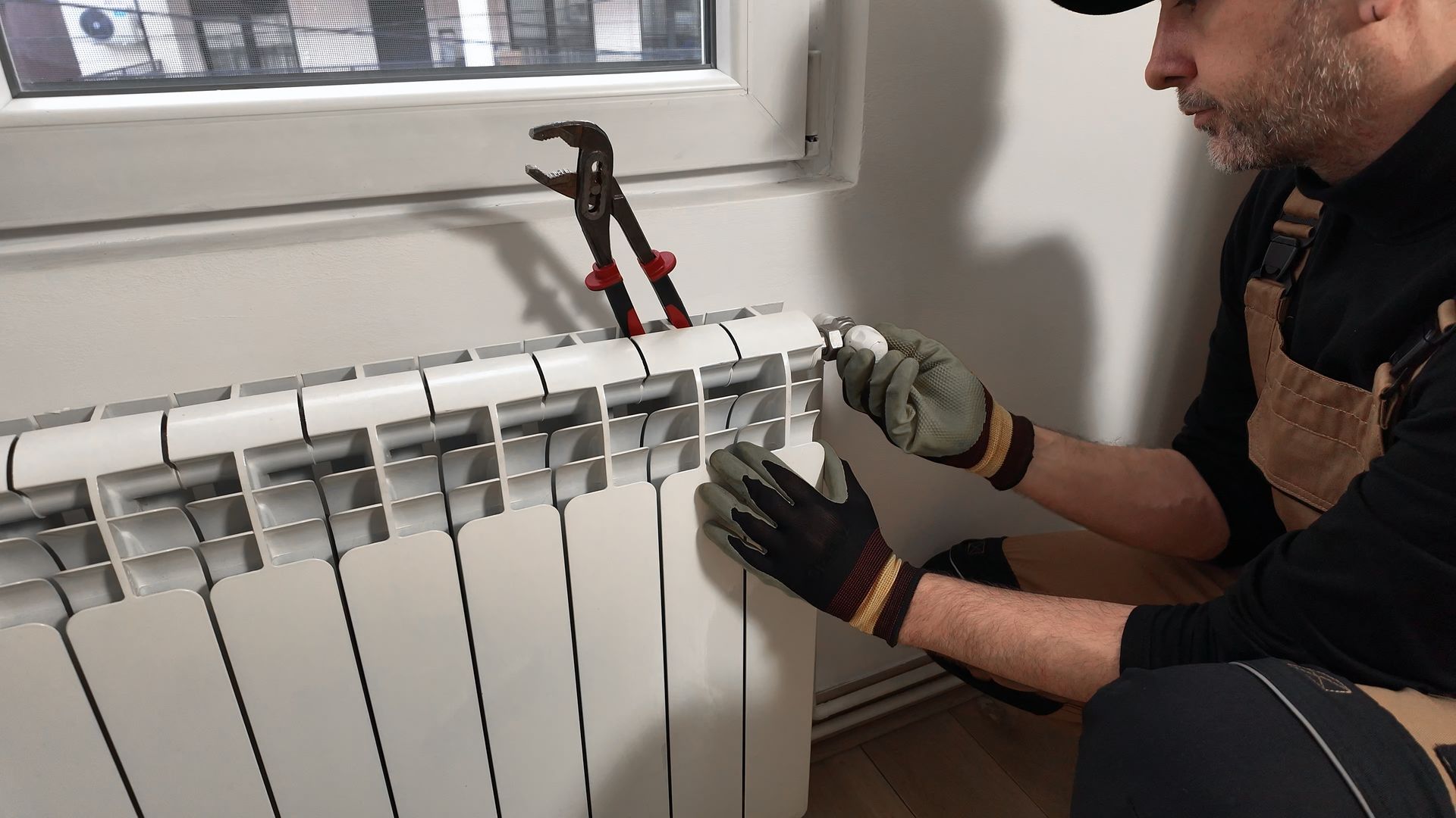 A person wearing work gloves adjusts the thermostat dial on a white radiator with pliers resting nearby.