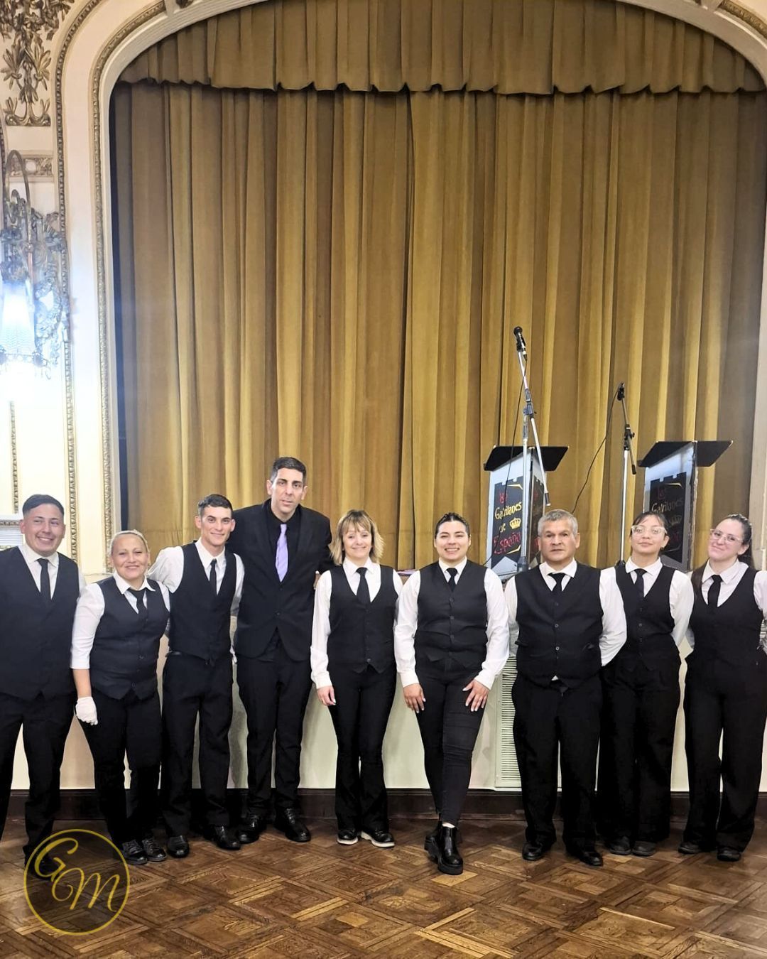 Grupo de personas con chalecos negros y camisas blancas posando frente a un escenario con una cortina dorada.