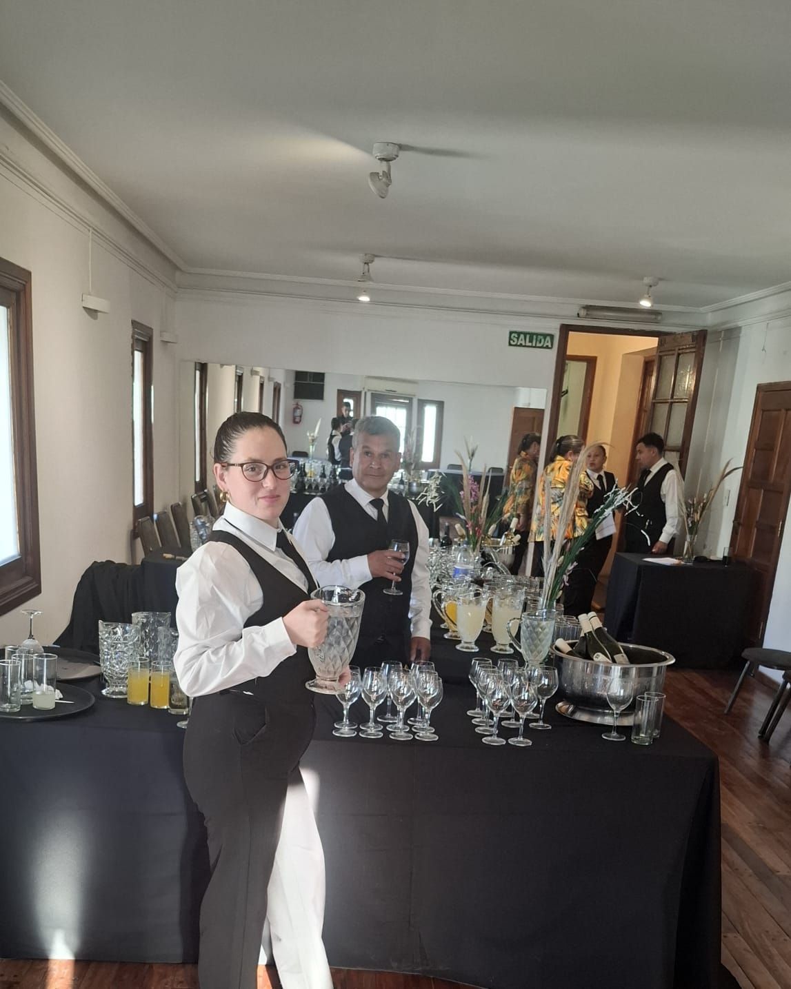 Dos camareros en una estación de bebidas. Uno sostiene una jarra y el otro un vaso. Camisas blancas y chalecos negros.