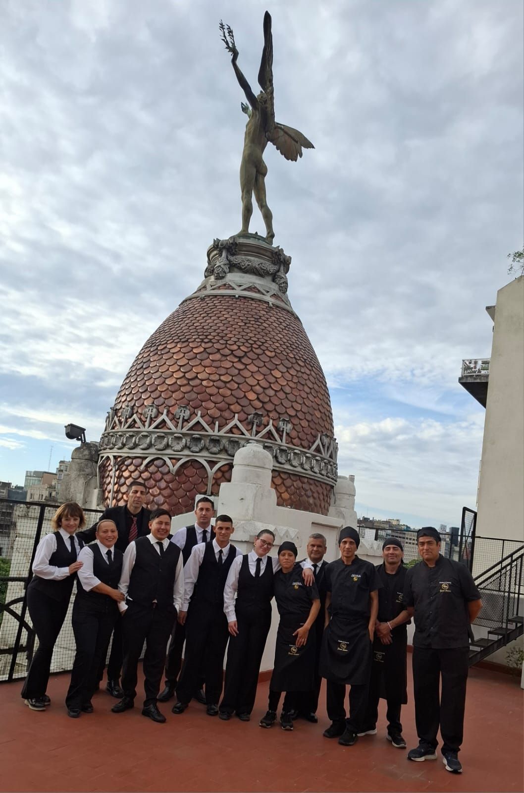 Grupo de personas uniformadas en una azotea, posando frente a una estatua encima de un edificio abovedado.