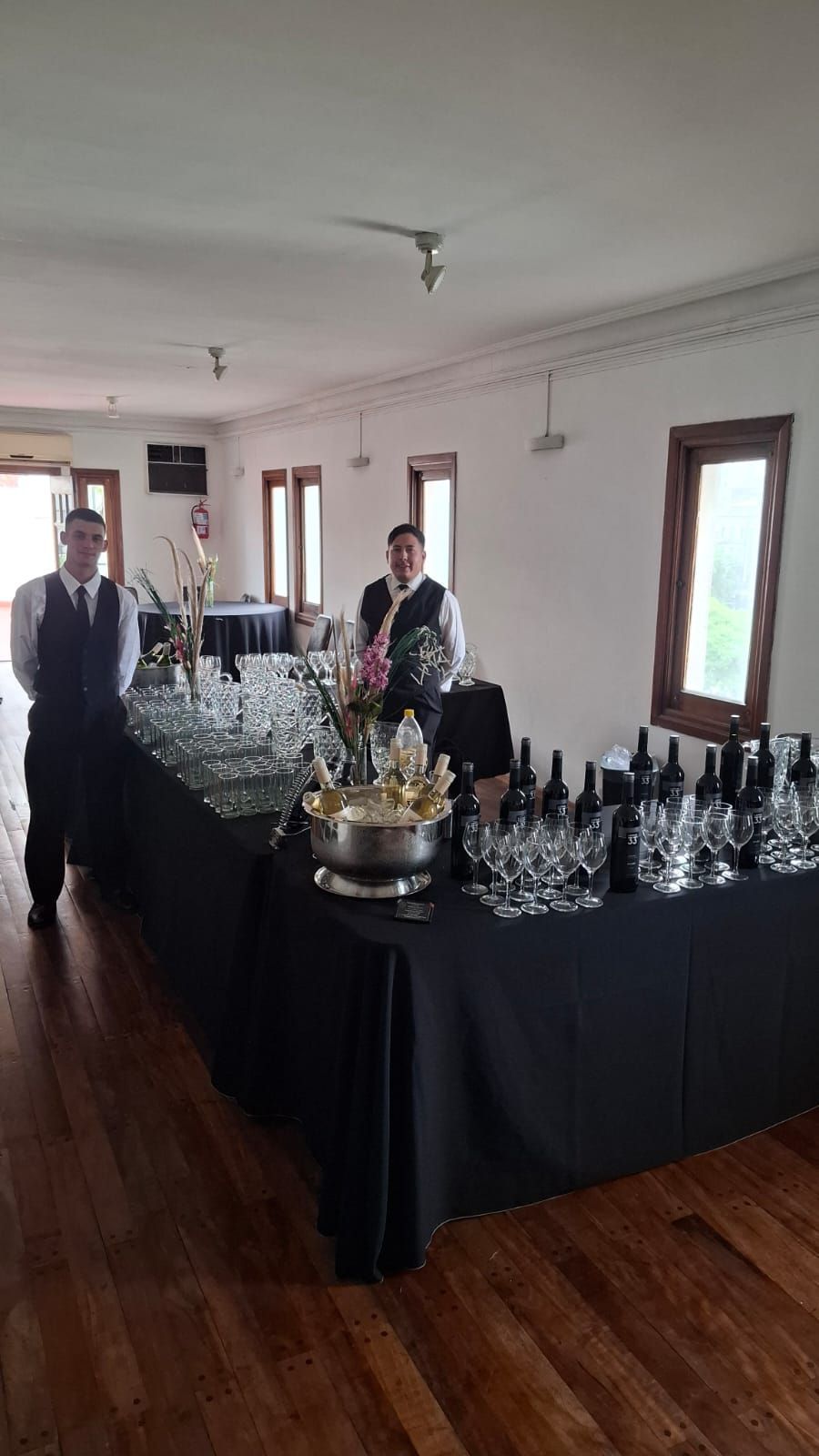 Dos camareros detrás de una mesa larga con vasos y botellas en un local con ventanas y suelos de madera.