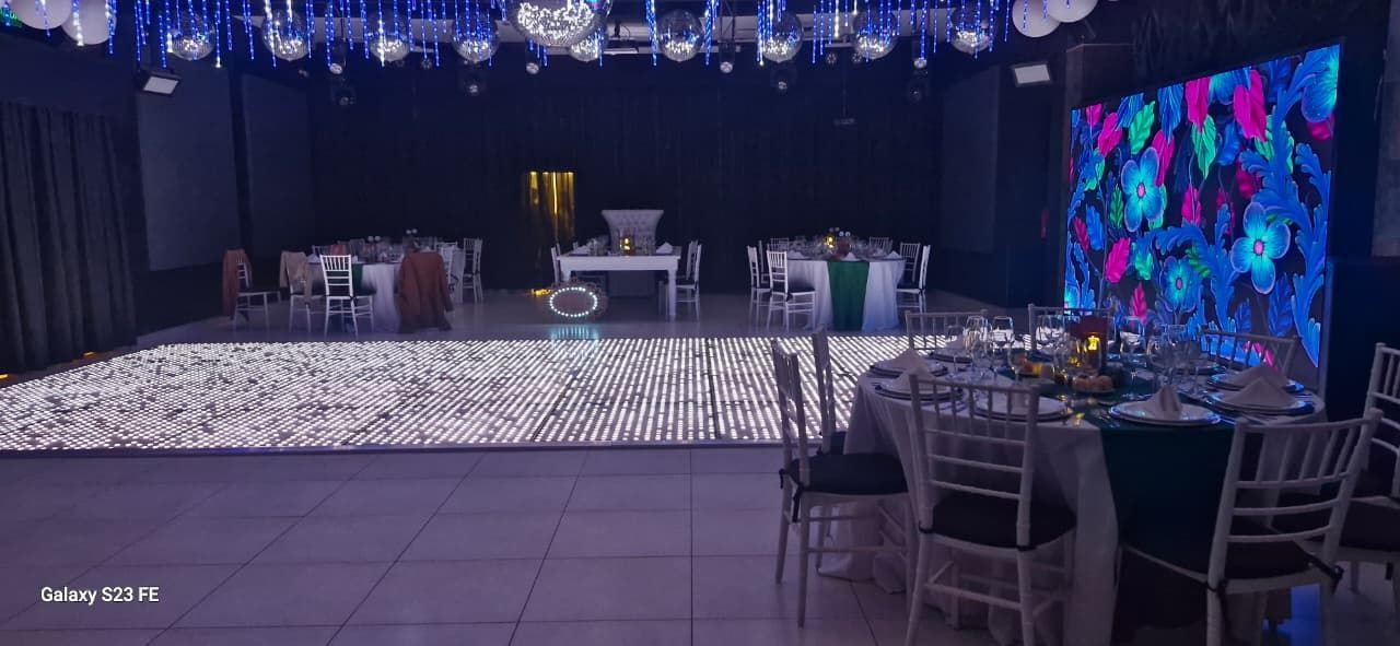Un salón de baile preparado para un evento, con mesas con cubiertos, una pista de baile iluminada y una pantalla colorida.