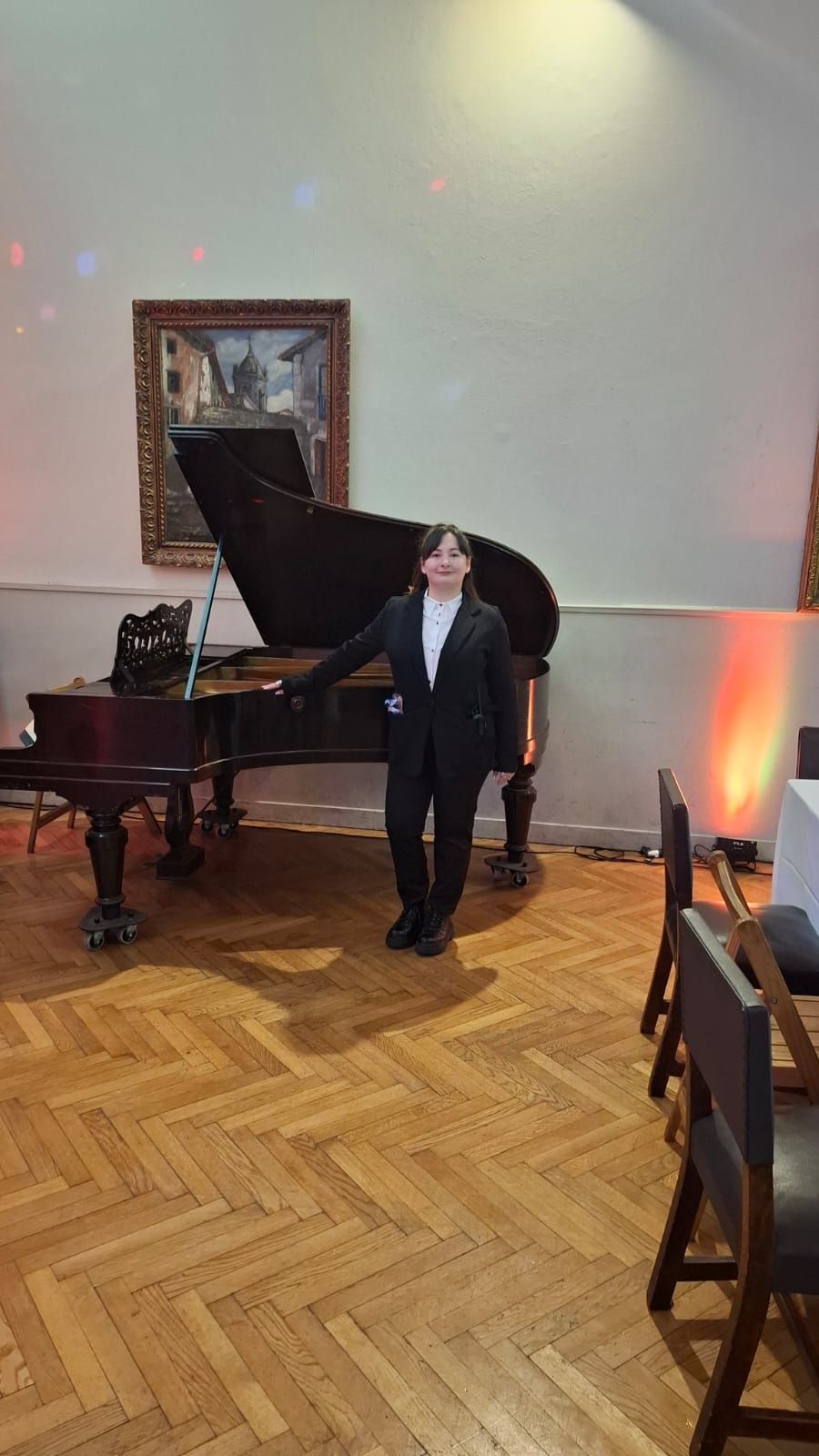 Una persona con traje se encuentra junto a un piano de cola en una habitación con un cuadro, sillas y suelo de parquet.