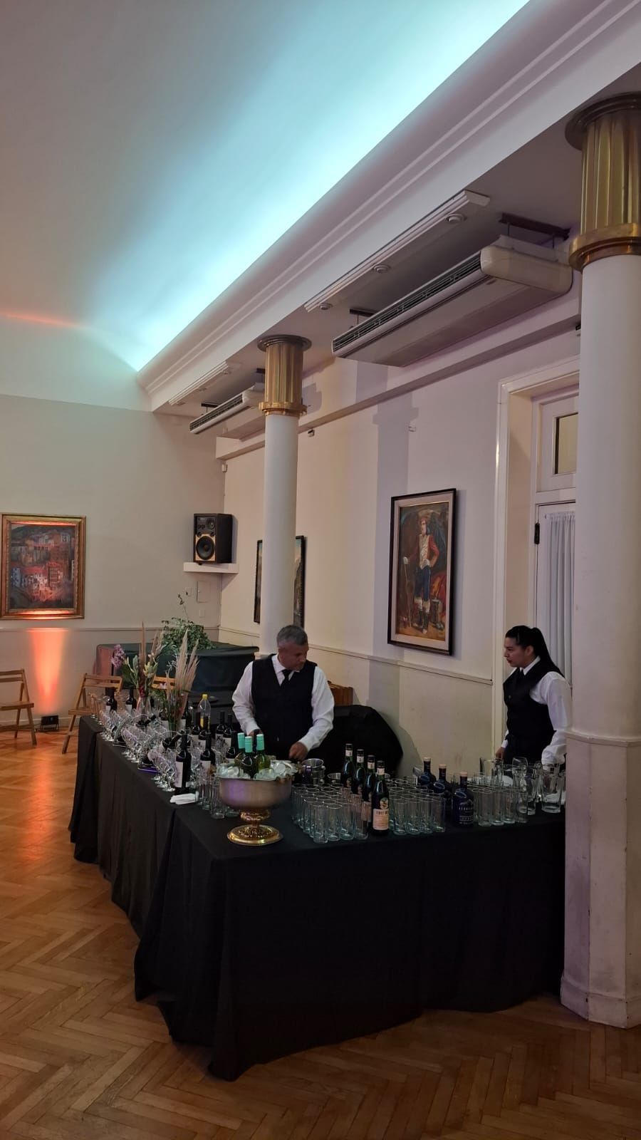 Barra con dos bartenders, botellas, cristalería, mesa negra y columnas blancas en un espacio para eventos.