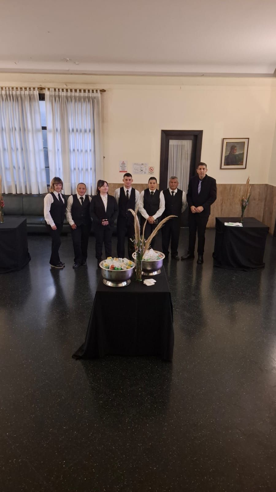 Grupo de camareros con uniformes negros, de pie en una habitación con dos mesas decoradas.