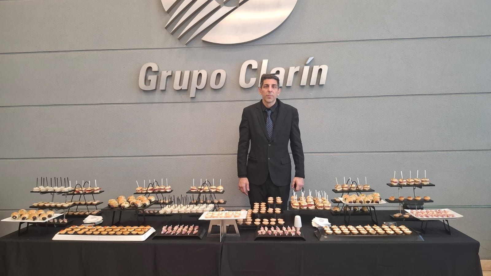 Un hombre de traje se encuentra detrás de una mesa repleta de aperitivos, frente al logo del