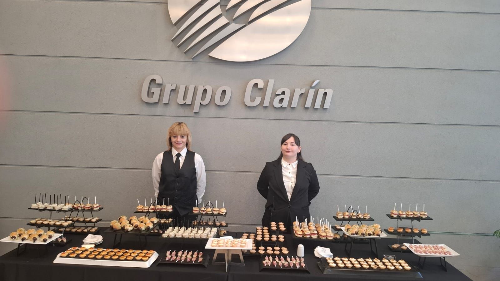 Dos personas permanecen detrás de un buffet de aperitivos frente a un cartel de