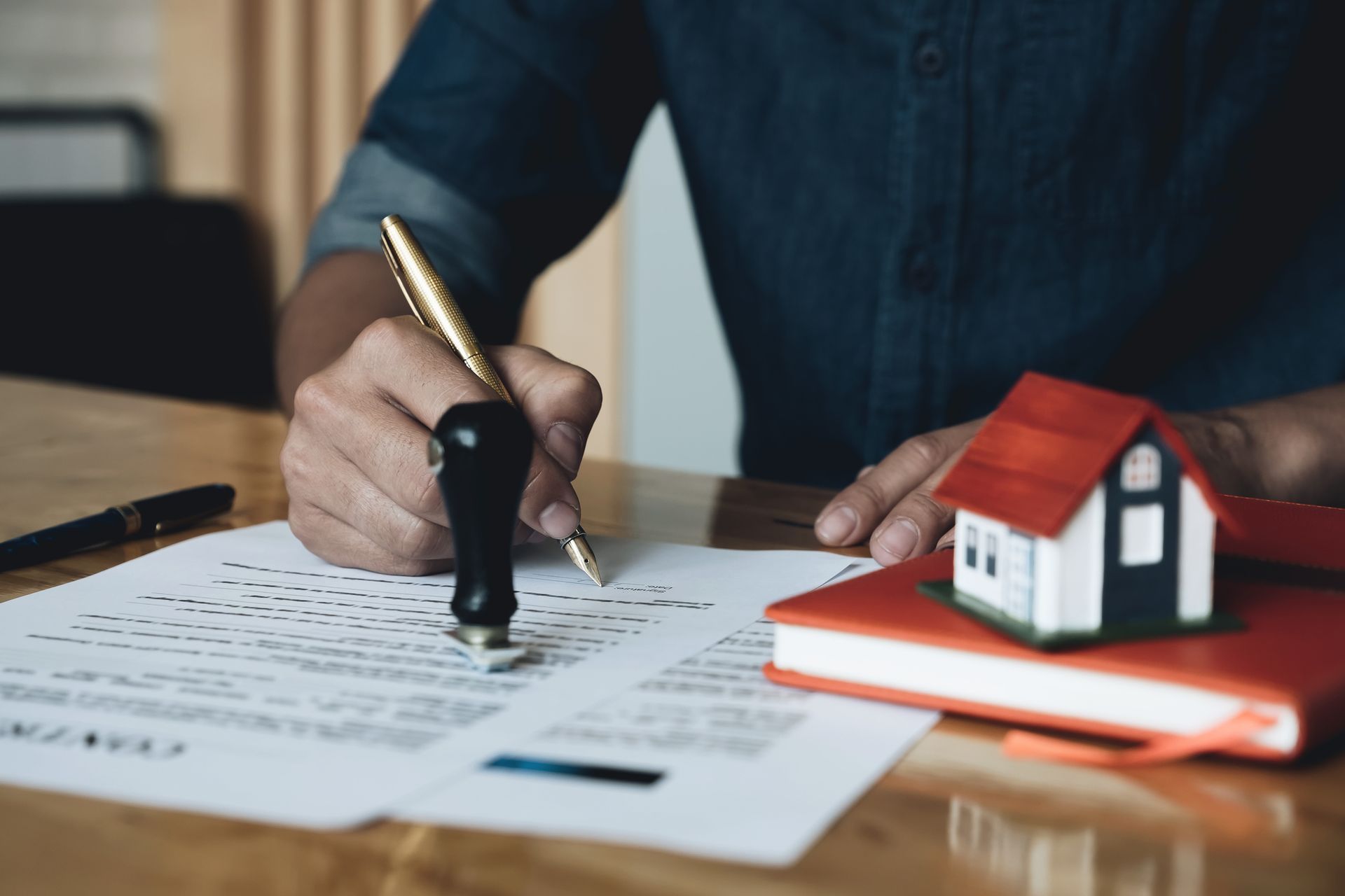 Person signing document with a pen and stamp next to a house model on a red book.