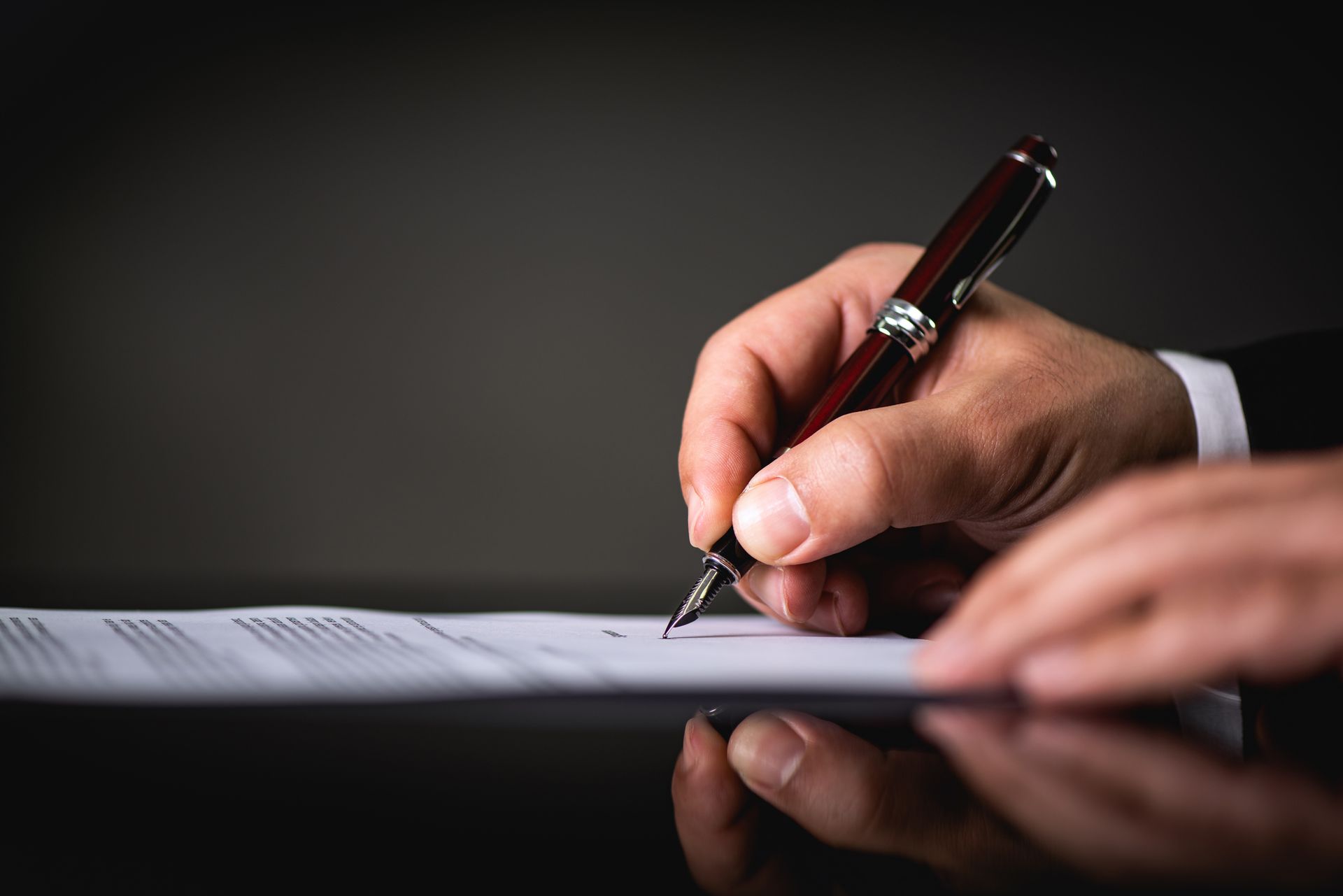 Hand writing on document with a pen; dark suit, sleek desk, focused.