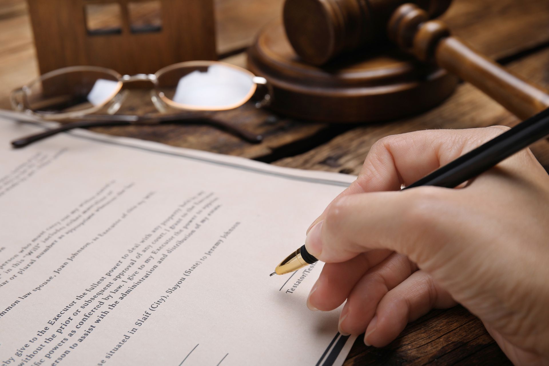 Hand signing a document with a fountain pen; gavel, glasses, and small house on a wooden table.