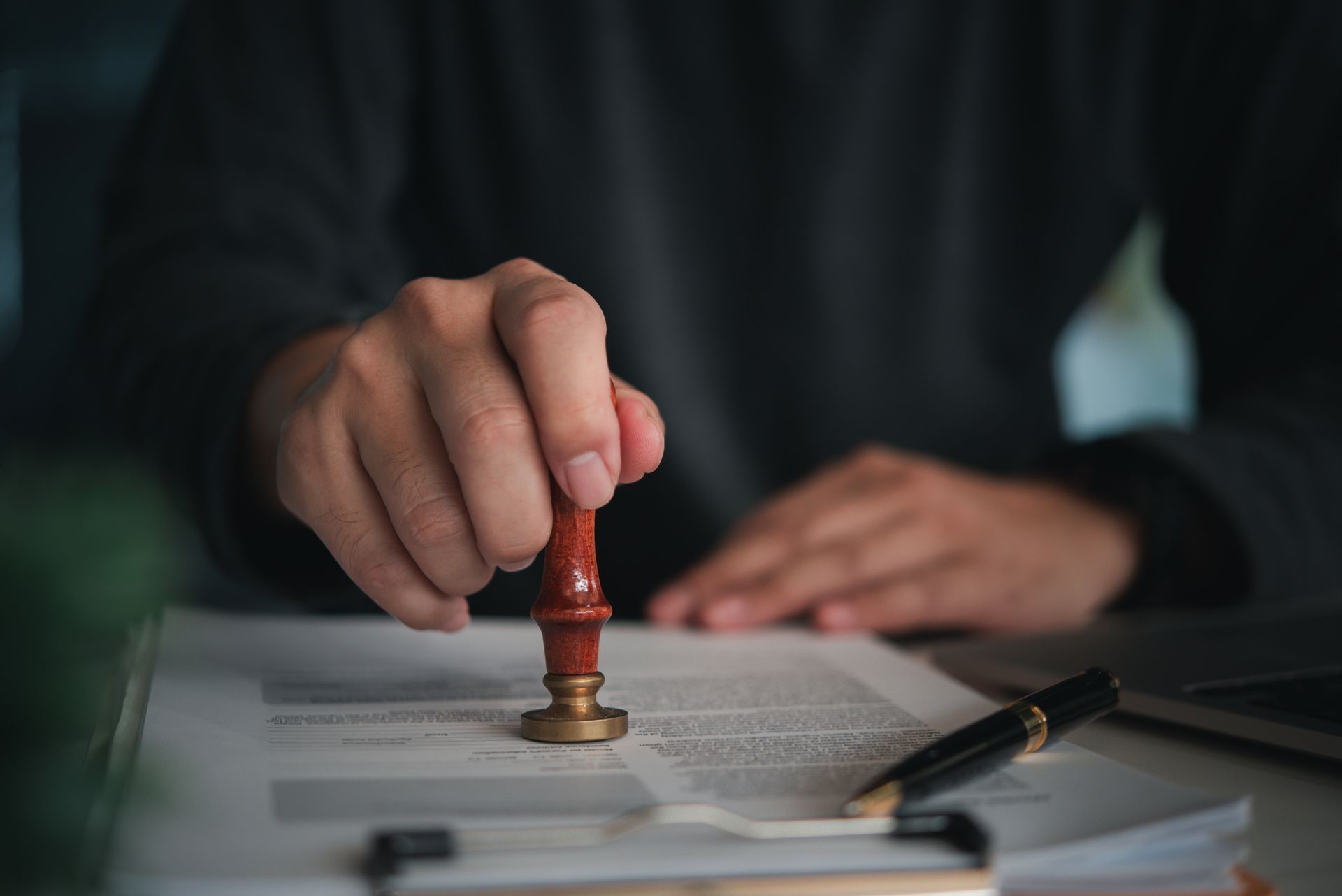 Person stamps a document with a wooden seal, pen and laptop visible.