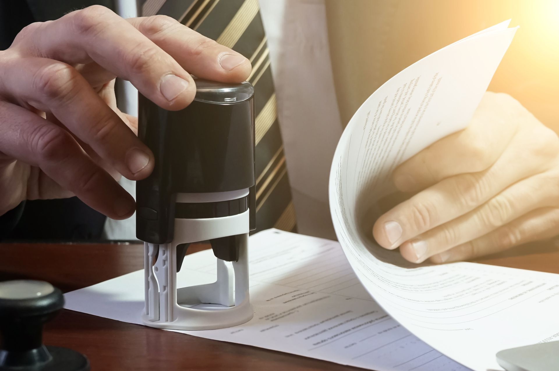 Person stamping a document with a black stamp on a desk, sunlit background.