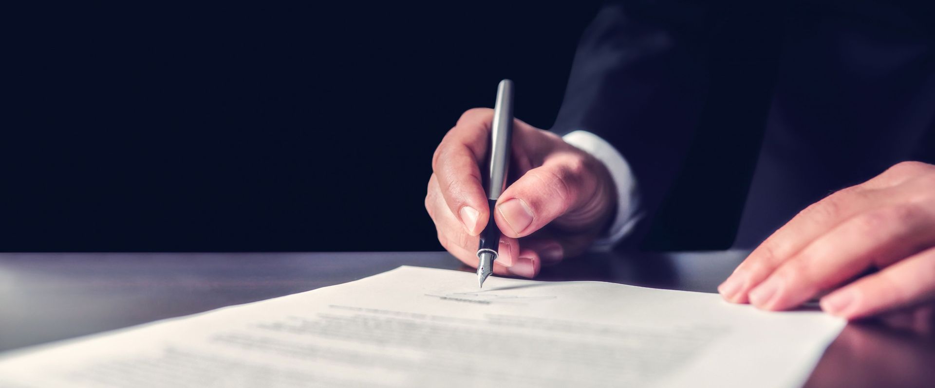 Person in a suit signing a document with a pen. Dark background.