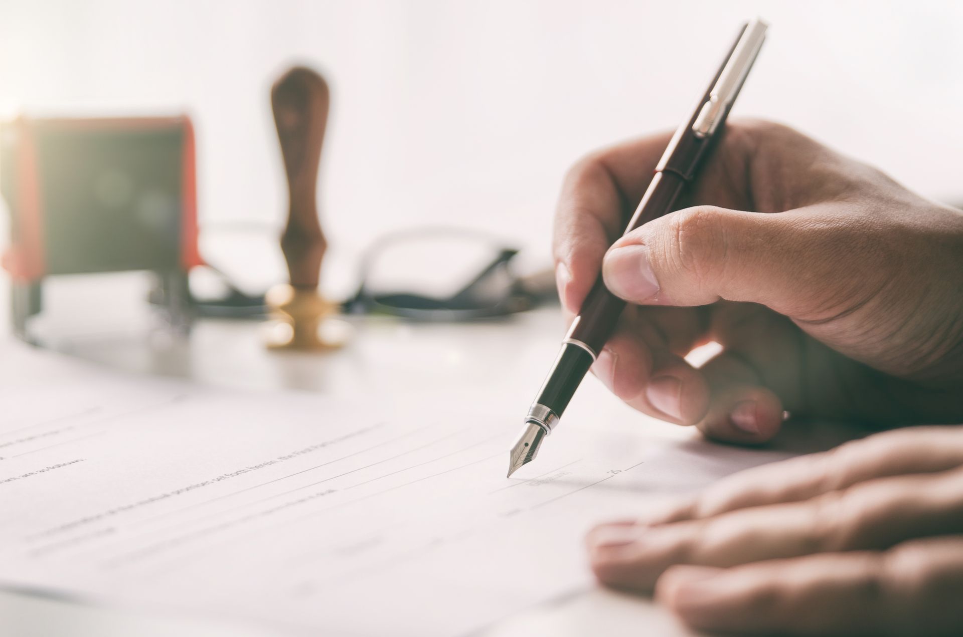 Hand signing document with a fountain pen; stamp and glasses in background.