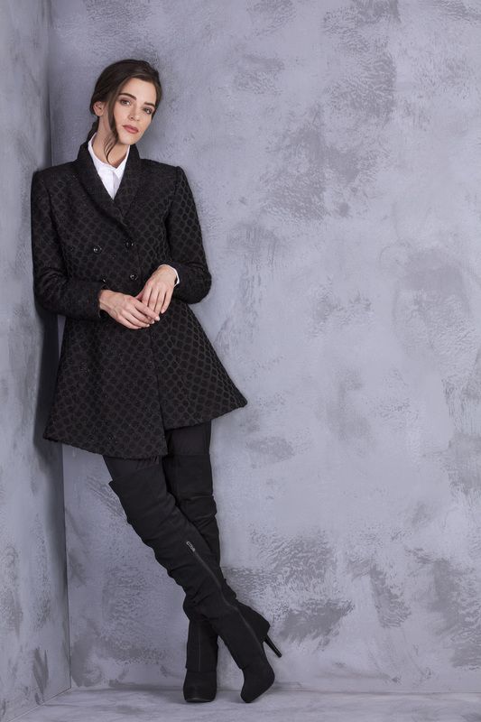 A woman in a black coat and boots is leaning against a wall.