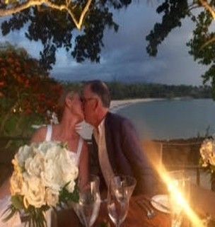 A bride and groom are kissing at a dinner table overlooking the ocean.