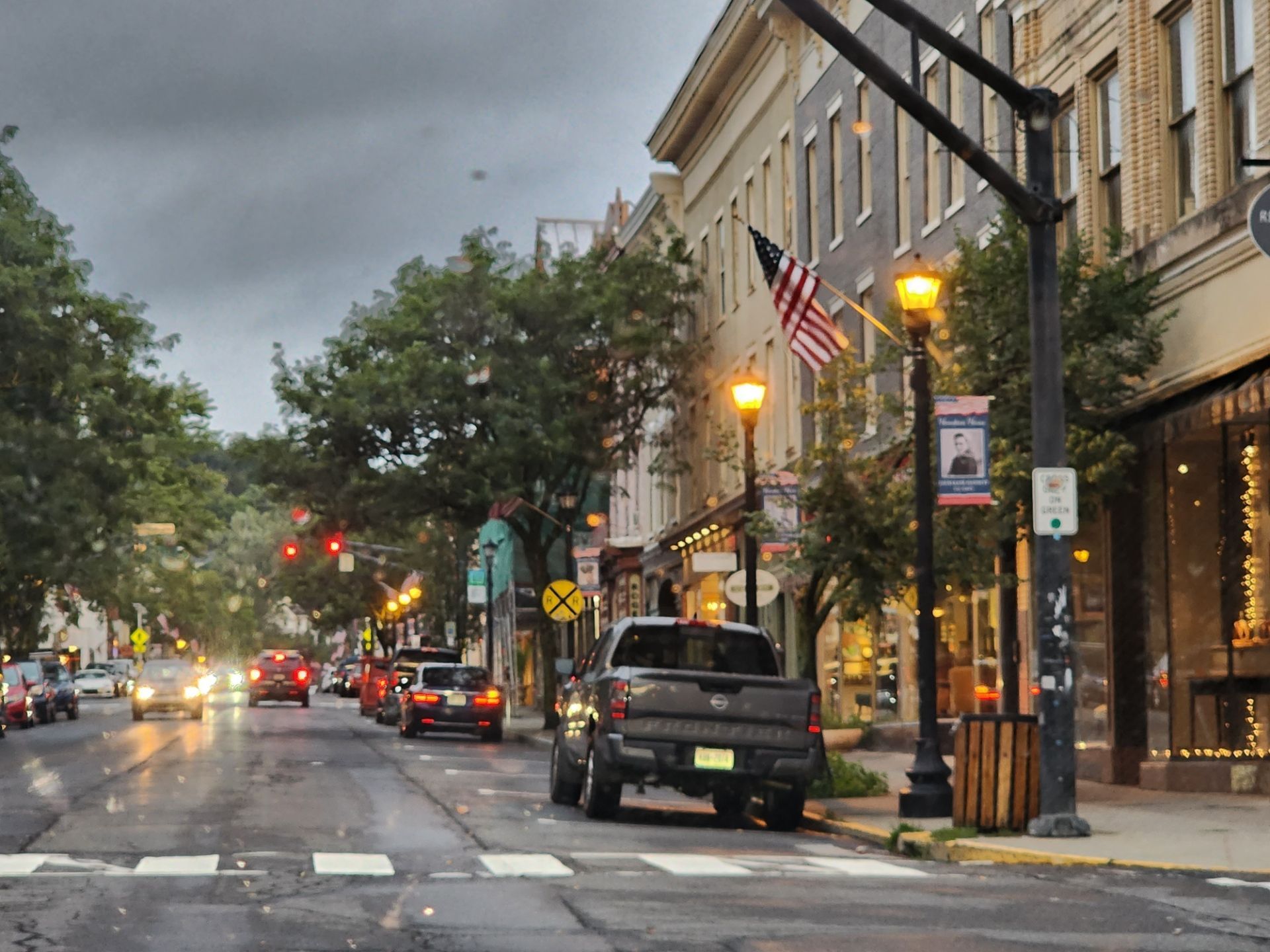 A wet street in a small town lined with shops, cars, and trees; a truck parked near a crosswalk.