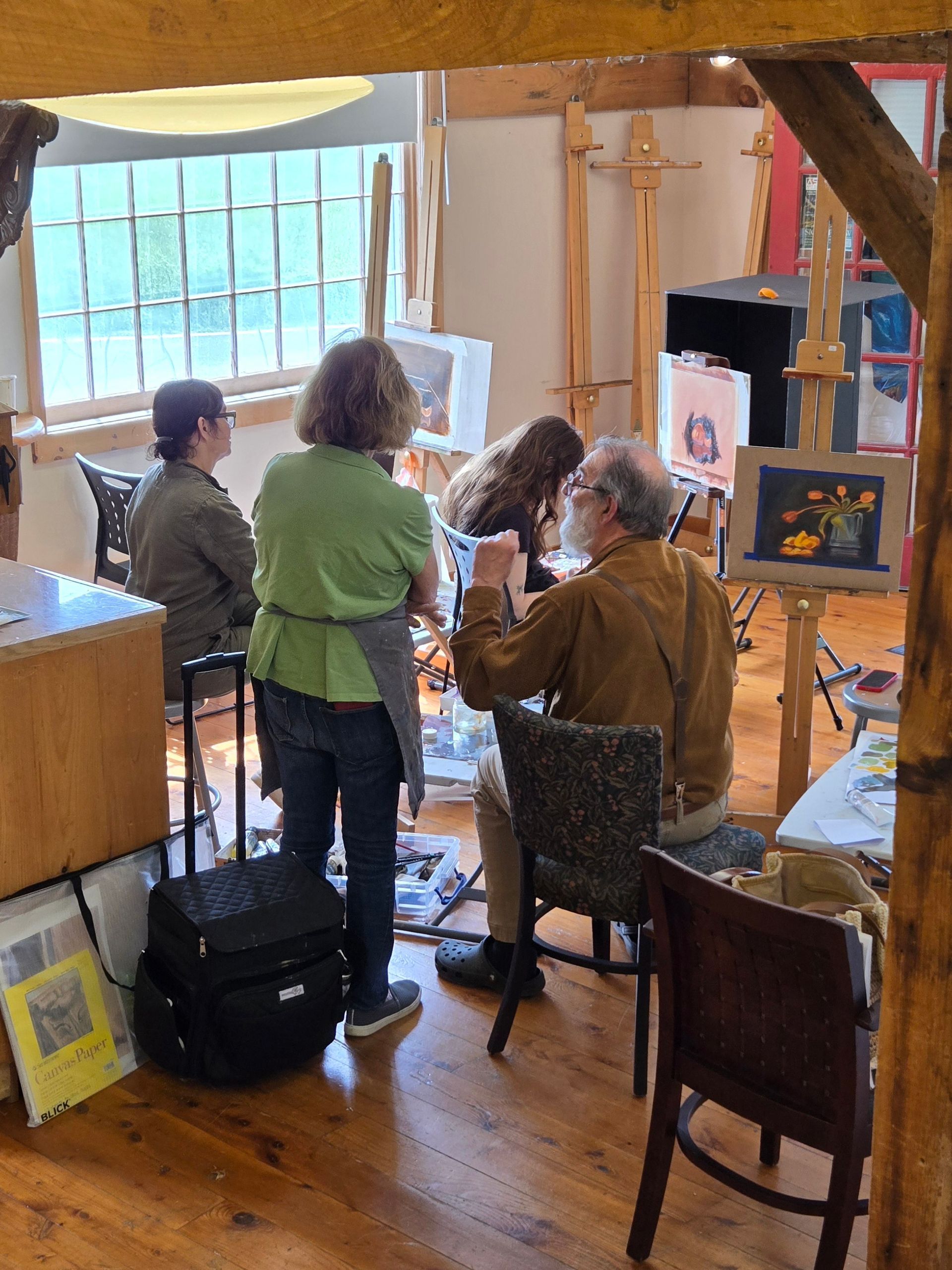 People in an art studio; a man painting while a woman assists. Other people watch in the background.