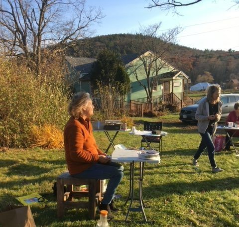People painting outdoors. Woman in orange shirt sits at table. Green house and mountain in background.