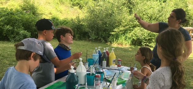 Children outdoors painting with assistance from an adult, surrounded by trees and colorful paint bottles.