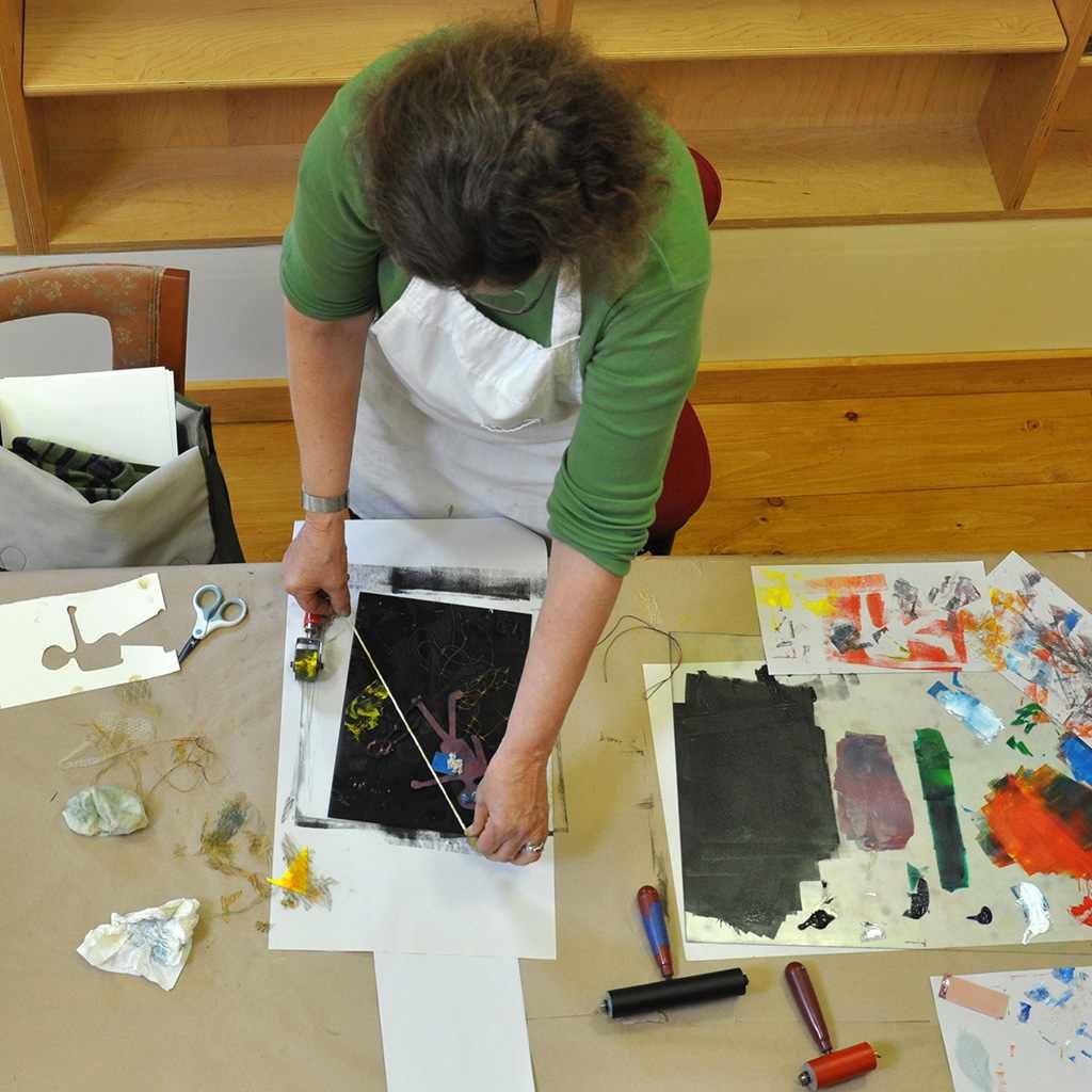 Person in green shirt and apron works on art print, using tools on a table in a studio setting.