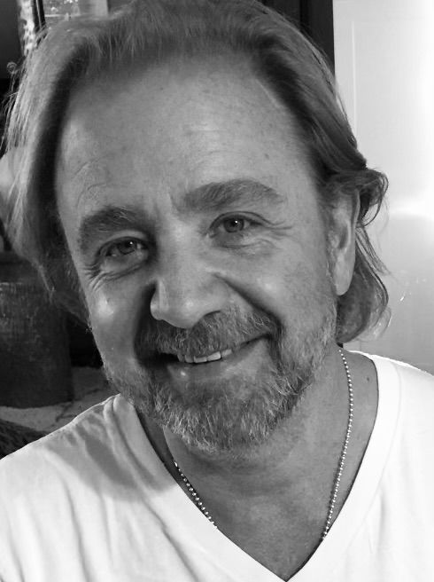 Smiling man with shoulder-length hair and beard, wearing a white shirt and necklace. Black and white.