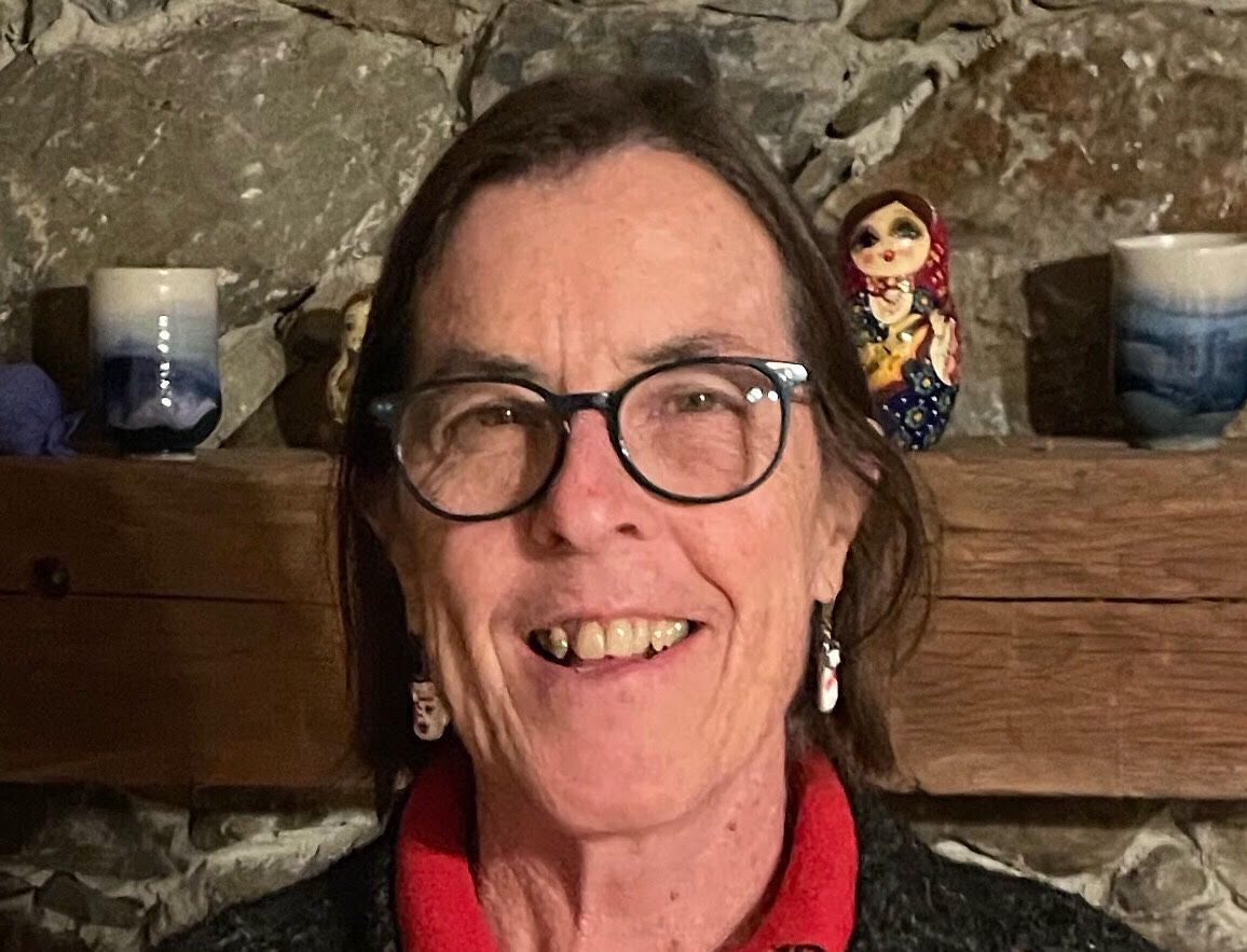 Woman with glasses smiles at the camera, red collar, earrings, stone fireplace background with decor.