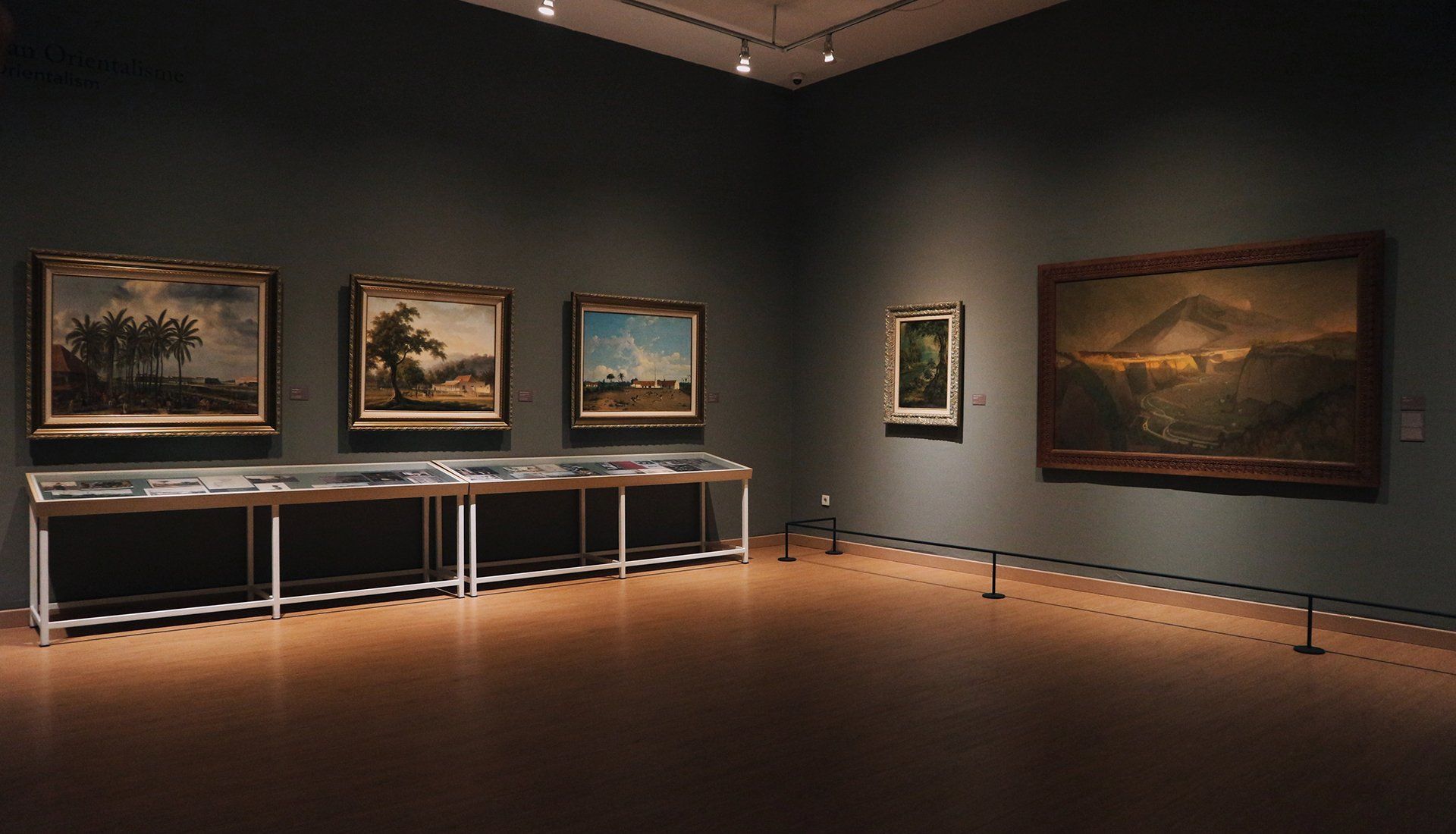 An art gallery room with landscape paintings hanging on a dark wall; display cases sit below.