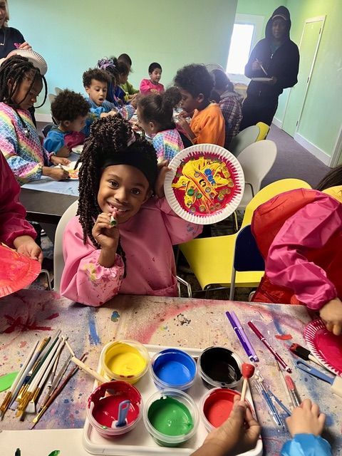 Children painting at a table. A girl smiles, holding up her colorful artwork. Paint, brushes, and colors are visible.