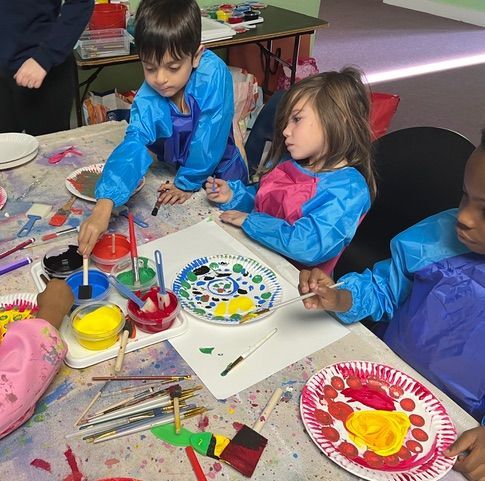 Children paint at a table, wearing smocks, with paint pots and paper plates.