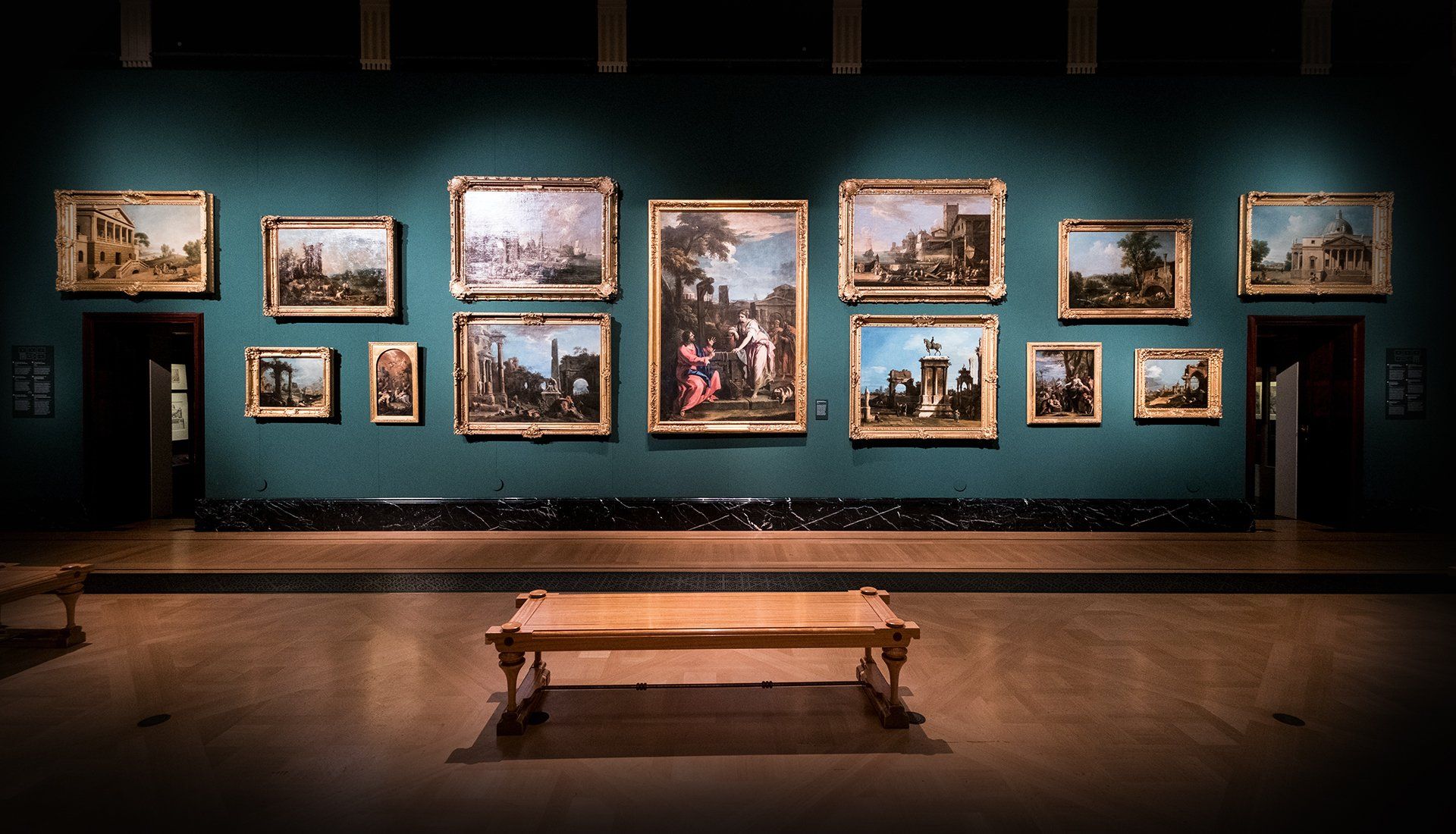 Art gallery with framed landscape paintings on a teal wall; a wooden bench sits below.