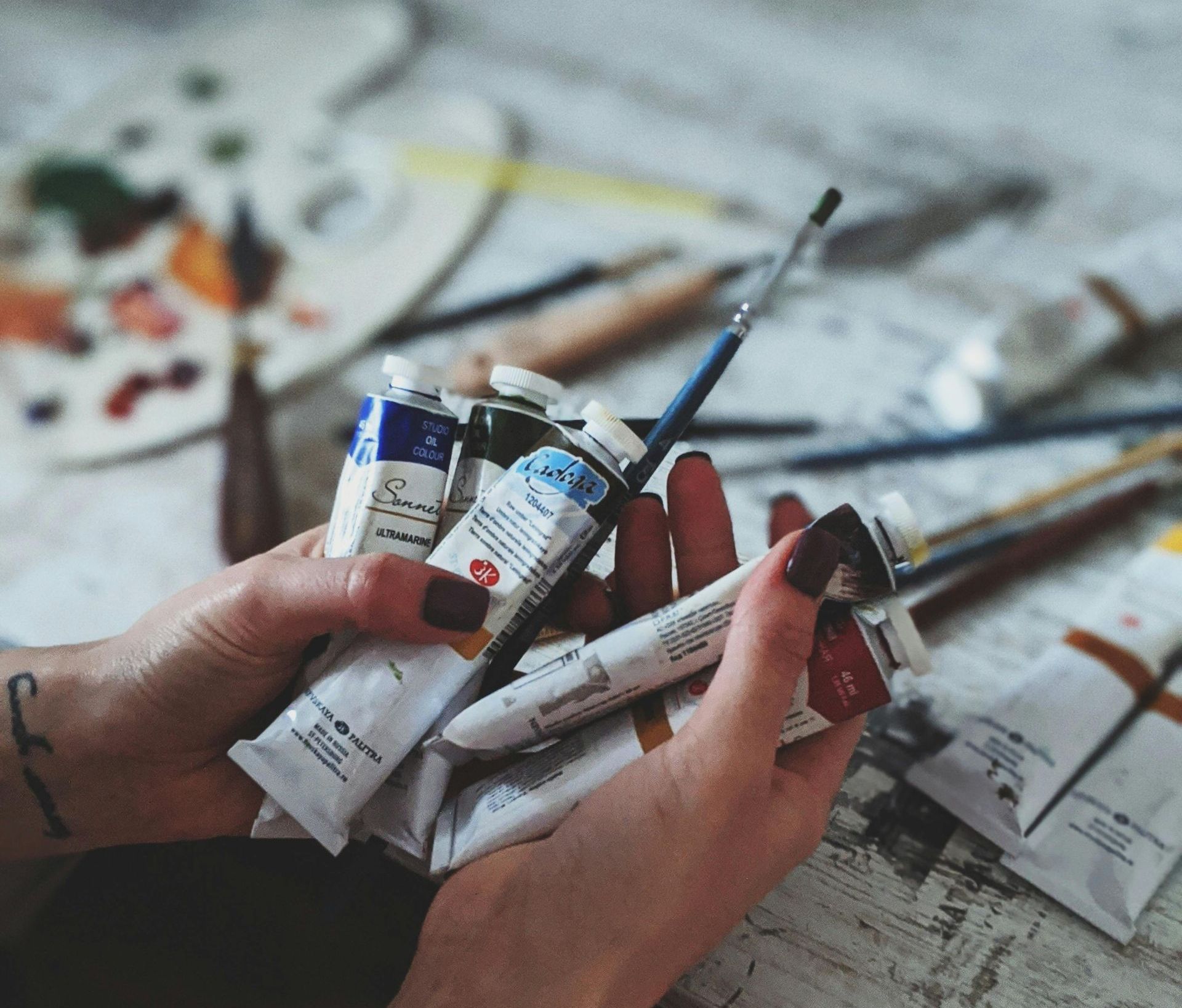 Hands holding tubes of paint with a paintbrush, palette, and other supplies in the background.