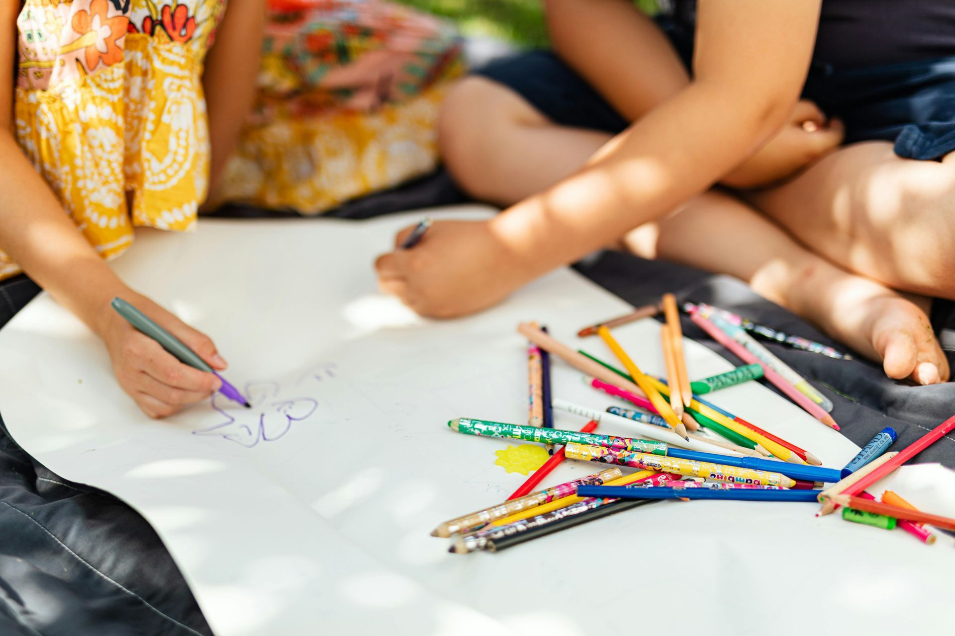 Two children drawing together on a large sheet of white paper, surrounded by colorful pencils, outdoors.