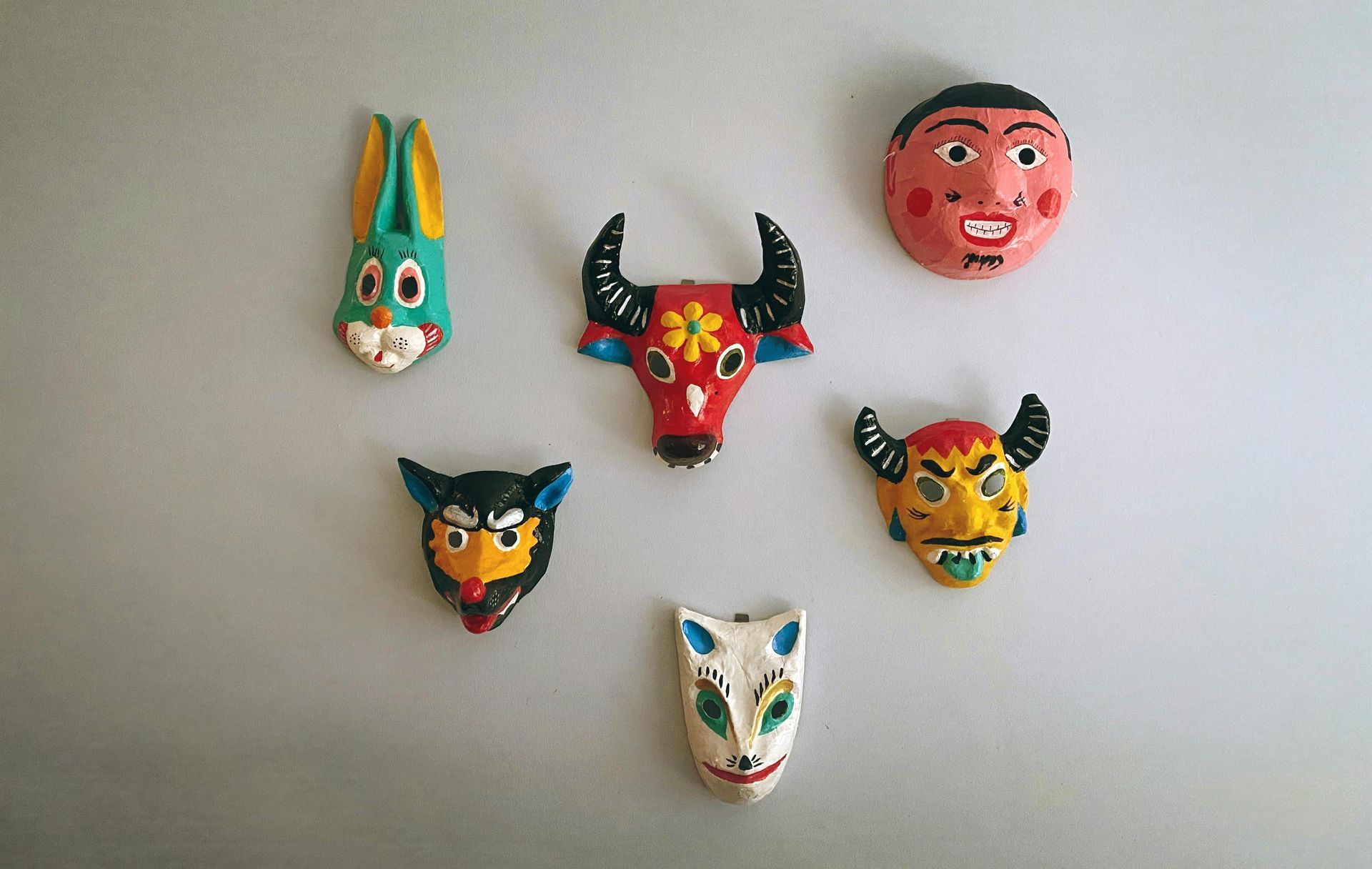 Six colorful, folk art masks hanging on a light gray wall: a bunny, bull, human face, demon, fox, and another demon.