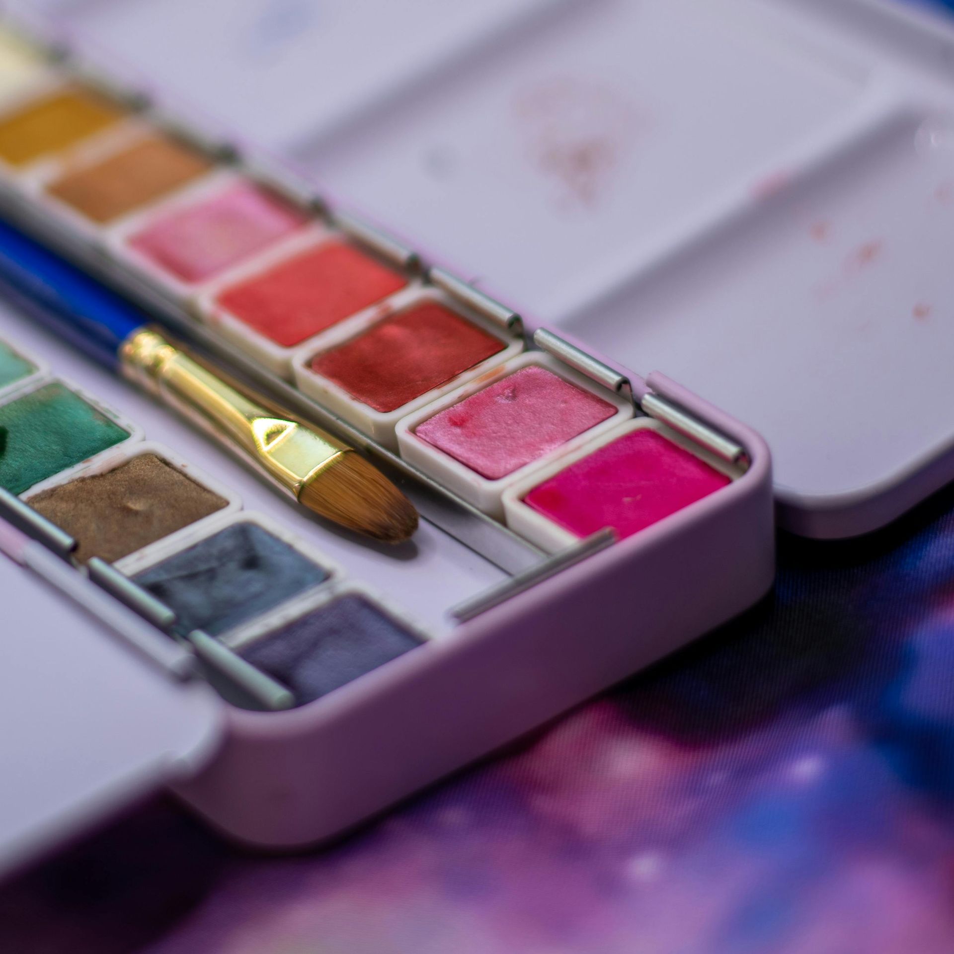 Watercolor paint palette with brush, various colors, purple case, galaxy background.