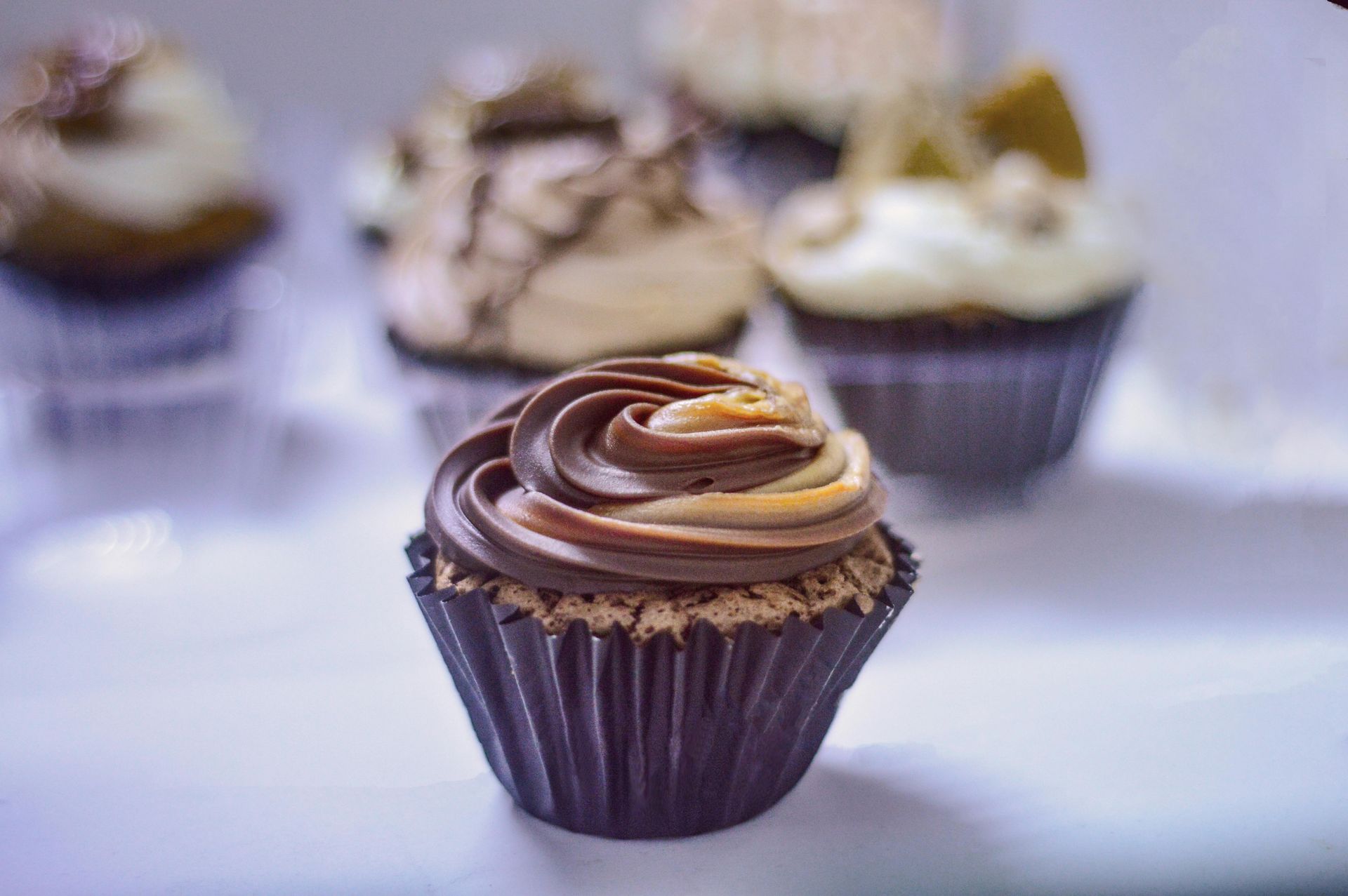 Chocolate cupcake with swirled frosting in a dark paper cup, surrounded by other frosted cupcakes on a white shelf.