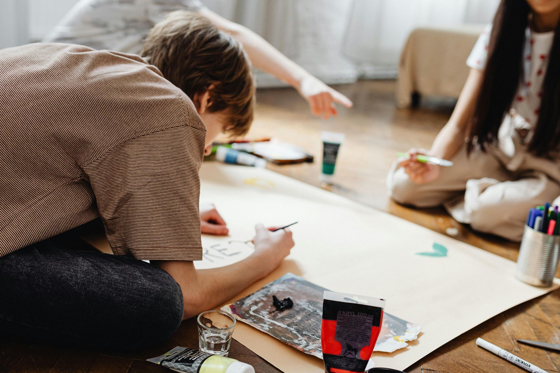 Three children on the floor painting on a large sheet of paper; paint tubes and brushes are visible.