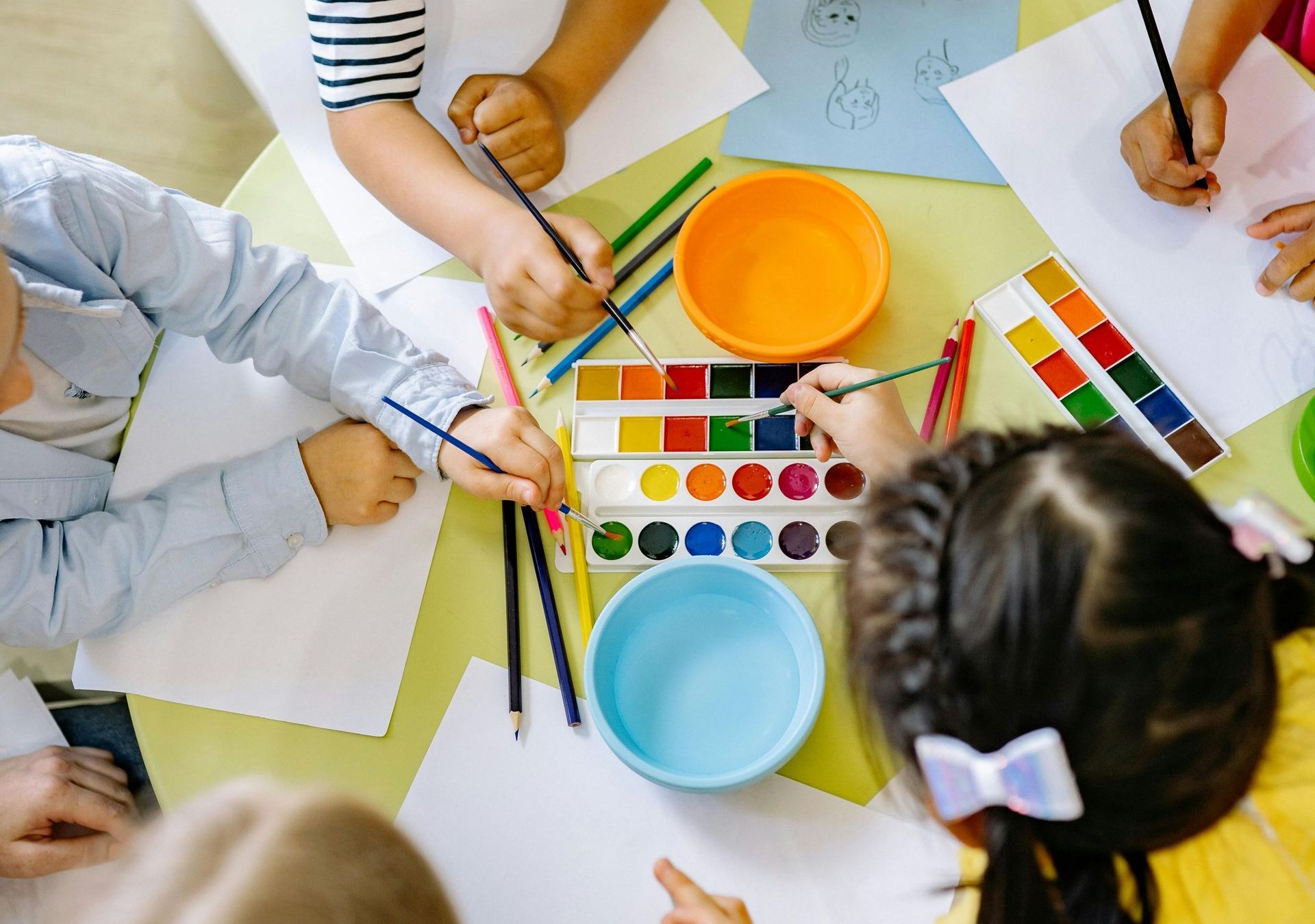 Children painting at a table: watercolor paints, brushes, paper, and colorful bowls.