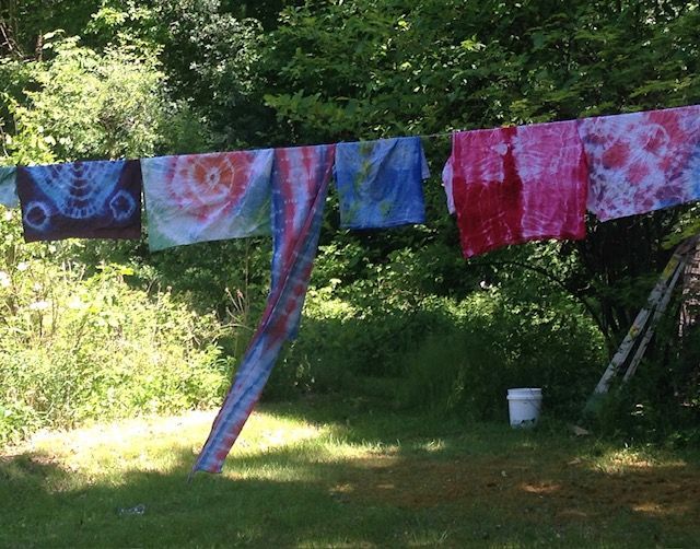 Tie-dye fabrics hang on a clothesline in a sunny, grassy outdoor setting.