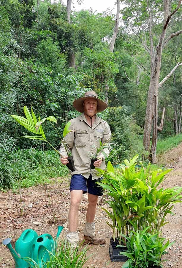 A Man is Standing Next to a Green Watering Can and Holding a Plant — Full Circle Landscaping in Alstonville, NSW
